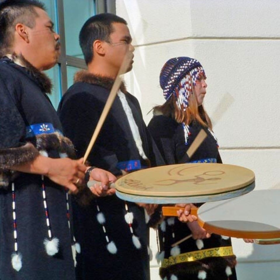 Three people beating large, circular, hand held drums.